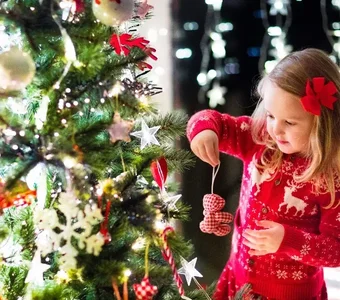 Blondes Mädchen mit rotem Weihnachtspulli schmückt den Christbaumbaum mit einem roten Teddyanhänger