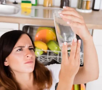 Frau vor einer Spülmaschine beäugt skeptisch ein Glas mit einem Grauschleier