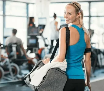 Eine Frau in sportlicher Kleidung steht in einem Fitnessstudio und trägt eine graue Sporttasche über der Schulter