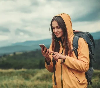 Eine junge Frau trägt einen Wanderrucksack und blickt in hügeliger Landschaft auf ihr Smartphone