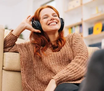Lächelnde junge Frau sitzt vor Couch und genießt Musik mit Over-Ear-Kopfhörern