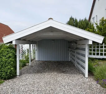 Weißes Holzcarport mit Satteldach steht auf einer Einfahrt aus Kieselsteinen