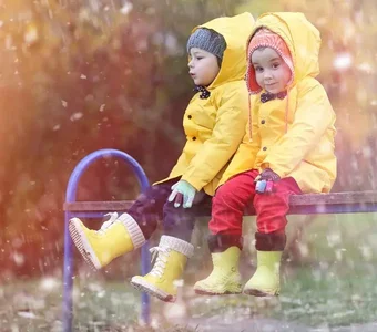 Junge und Mädchen mit gelben Regenjacken und Gummistiefeln sitzen im Regen auf einer Bank