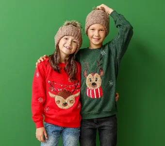 Ein Junge und ein Mädchen mit beigen Strickmützen und roten und grünen Pullovern mit weihnachtlichen Rentiermotiven vor grünem Hintergrund