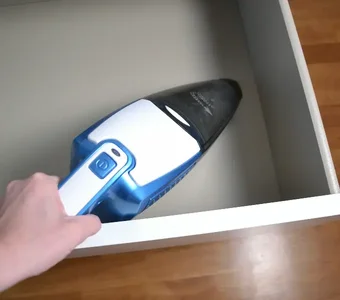 Eine Person saugt mit einem weiß-blauen Handstaubsauger in der Schublade eines weißen Schranks