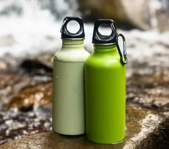 Zwei Metall-Trinkflaschen in grün und beige stehen nebeneinander auf nassem Boden