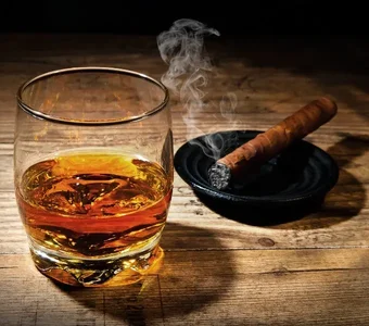 Ein Glas mit braunem Rumsteht auf einem Holztisch neben einer kubanischen Zigarre