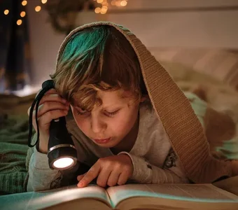 Junge mit einer Wolldecke über dem Kopf liest mithilfe einer Taschenlampe ein Buch