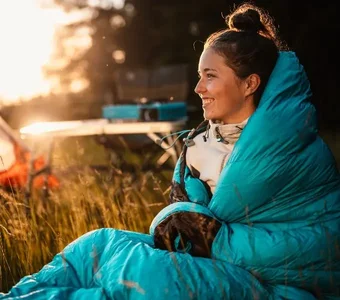 Junge Frau mit türkisfarbenem Daunenschlafsack sitzt draußen in der Natur