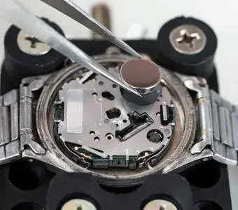 Abbildung einer geöffneten Armbanduhr, bei welcher gerade die Batterie gewechselt wird.