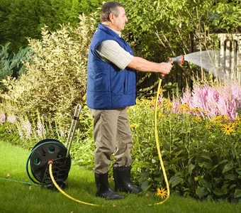 Mann, der mit einem auf einem Schlauchwagen aufgerollten Gartenschlauch Gartenpflanzen bewässert