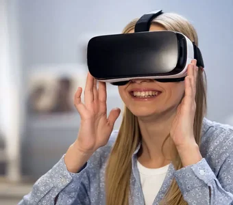 Aufnahme einer jungen Frau, die eine VR-Brille im Gesicht trägt, begeistert guckt und mit beiden Händen an die Außenkanten der Brille fasst