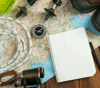Auf einem Holztisch liegen verschiedene Campingutensilien wie eine Landkarte, ein Kompass, Wanderschuhe oder ein Fernglas
