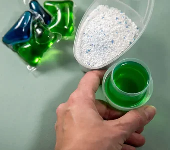 Vor einem hellgrünen Hintergrund befinden sich zwei Waschkapseln, Waschpulver in einer Dosierhilfe und flüssiges Waschmittel in einem Deckel, welches von einer Person gegriffen wird
