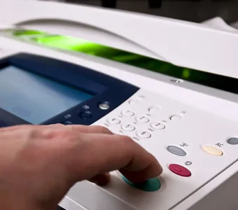Eine Hand bedient die Tasten eines Multifunktionsdruckers, im Hintergrund leuchtet grünes Licht unter der Scannerklappe des Geräts