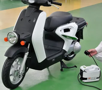 Schwarz-weißer E-Roller steht auf grünem Boden, Mensch schließt Ladegerät an das Fahrzeug an