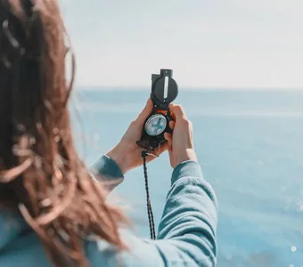 Frau am Berghang ermittelt die richtige Richtung mit einem Kompass - im Hintergrund ist das blaue Meer zu sehen