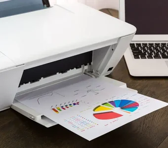 Ein Laserdrucker mit einem farbig bedruckten Dokument und ein Laptop stehen auf einem Holztisch