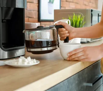 Nahaufnahme einer jungen Frau,  die eine Kaffeekanne aus der Kaffeemaschine nimmt, um einen Kaffee einzuschenken