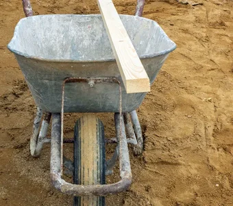 Eine Schubkarre aus Metall steht auf einem sandigen Untergrund. Eine Holzlatte liegt auf ihrer Mulde