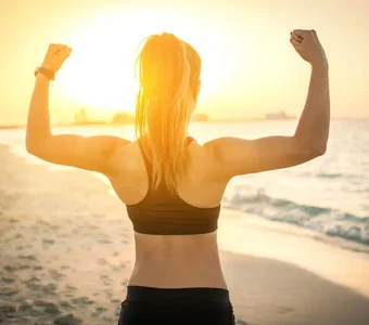 Eine sportliche Frau steht vor einem Sonnenuntergang am Strand und zeigt ihre Armmuskeln
