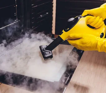 Eine Person mit gelben Gummihandschuhen reinigt einen Backofen mit einer Handdüse durch Dampf