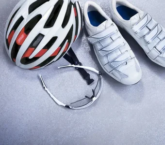 Auf einem grauen Untergrund befinden sich weiße Fahrradschuhe, eine Sportbrille und ein Schutzhelm