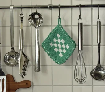 Ein grüner gehäkelter Topflappen hängt dekorativ in der Küche neben einigen Küchenutensilien