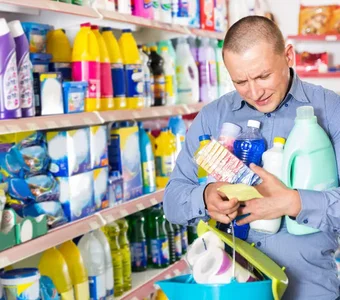 Mann steht in einem Laden vor Regalen voller Putzmittel, hält einige Reiniger in den Armen und schaut auf eine Liste