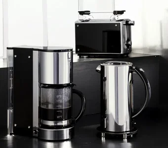Auf einer dunklen Arbeitsfläche befinden sich eine Kaffeemaschine, ein Wasserkocher und ein Toaster im Edelstahl-Design, welche zudem einige Kunststoffelemente aufweisen