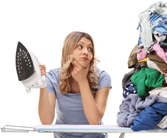 Blonde Frau schaut nachdenklich auf einen Wäscheberg und hält ein Bügeleisen in der Hand
