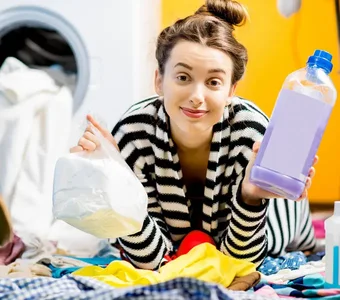 Junge Frau liegt zwischen ihrer Wäsche und hält Flüssig- und Pulverwaschmittel in der Hand