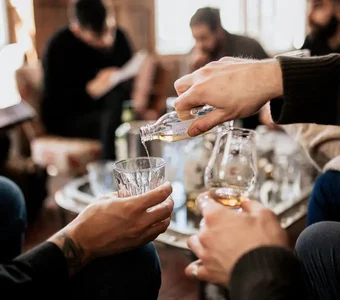 Eine Gruppe Personen sitzt um einen Tisch, sie schenken sich Whisky in ihre Gläser ein