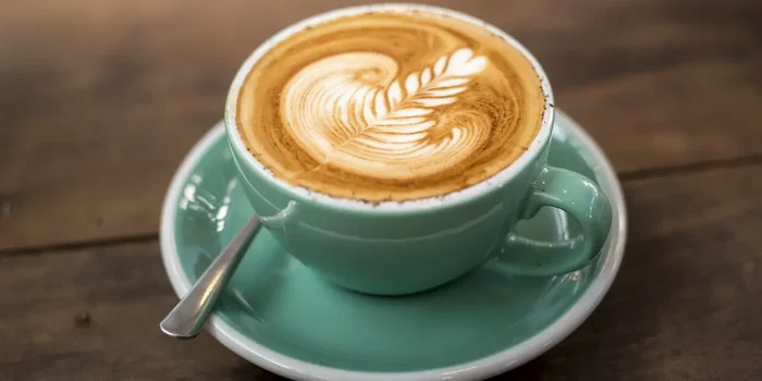 Cappuccino mit einem Blattmuster nach Latte-Art in einer mintgrünen Tasse steht auf einem dunklen Holztisch