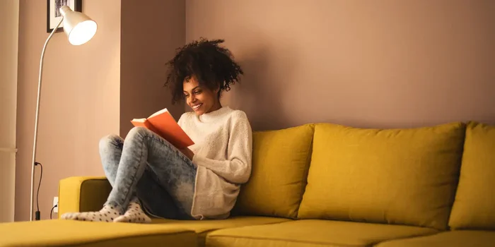 Eine Frau sitzt im Licht einer weißen Stehlampe auf einem gelben Sofa und liest ein Buch