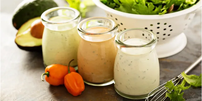 Gläser mit drei verschiedenen Dressings stehen vor einem mit frischem Salat gefüllten Küchensieb