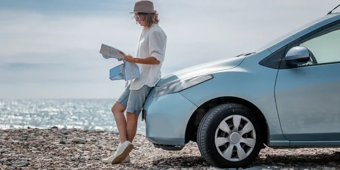 Eine Frau schaut in eine Landkarte, während sie an der Motorhaube eines silbernen Autos lehnt, das an einem Steinstrand steht