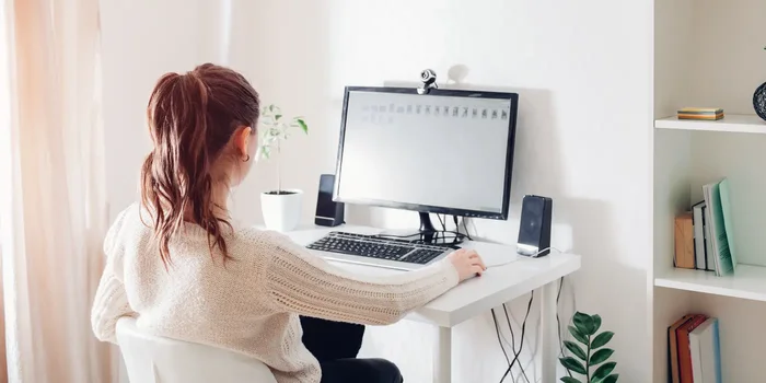 Frau sitzt an Schreibtisch mit PC, Lautsprechern und Webcam in hellem Büro