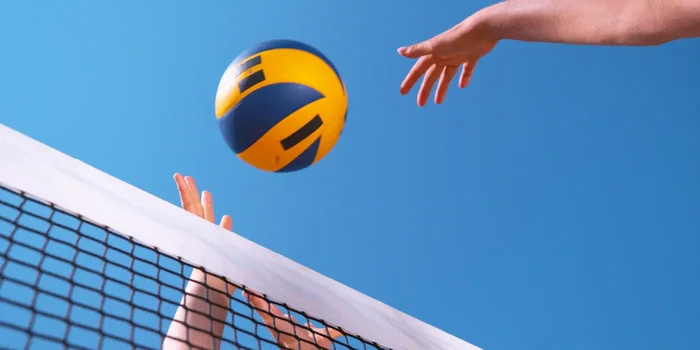 Ein blau-gelber Volleyball fliegt übers Netz, dabei sind ein blauer Himmel und die Hände der Spieler:innen zu sehen