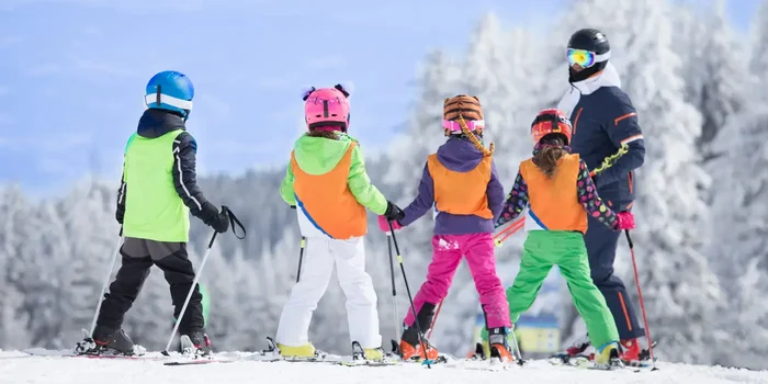 Vier bunt gekleidete Kinder mit Helm, Skistöcken und Ski hören draußen im Schnee einem Skilehrer zu