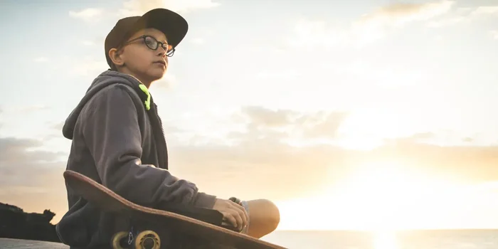 Ein Junge mit Cap, Brille und Kapuzenpullover sitzt bei Sonnenuntergang mit seinem Skateboard am Meer