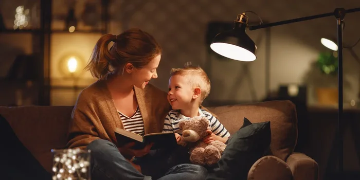 Eine Frau und ein kleiner Junge sitzen abends beim Licht einer Leselampe gemütlich auf einem Sofa und lesen gemeinsam ein Buch