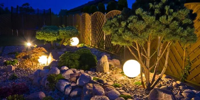 Ein Garten mit Steinen, Sträuchern und einem Holzzaun ist von kugelförmigen Lichtern erhellt