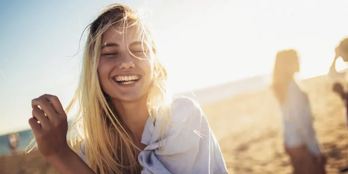 Eine junge Frau mit langen Haaren lacht am Strand in der Sonne
