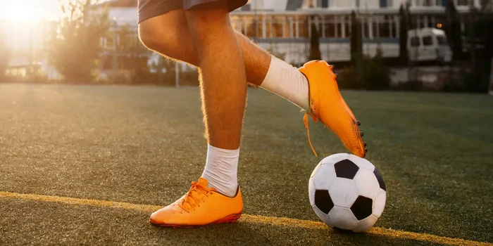 Model trägt einen orangefarbenen Nike Fußballschuh auf einem Kunstrasenplatz
