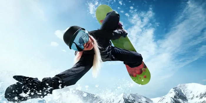 Frau mit schwarzer Snowboardkleidung und Brille beim Sprung mit einem grünen Snowboard