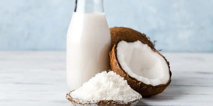 Vor einem hellen Hintergrund befinden sich eine Glasflasche mit Kokosmilch, Kokosmehl auf einem Teller und eine aufgeschlagene Kokosnuss