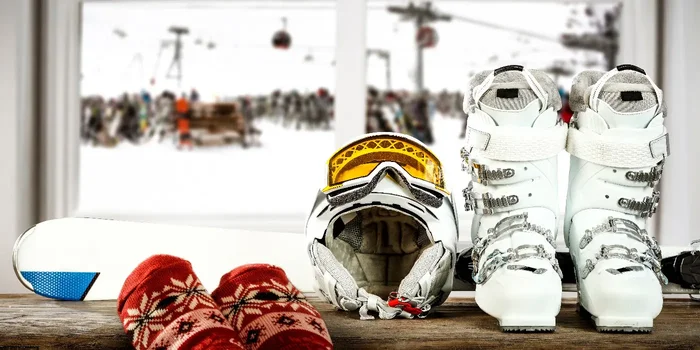Auf einem Fensterbrett mit verschneiter Landschaft im Hintergrund liegt eine weiße Skiausrüstung bestehend aus Helm, Schuhen und Skiern