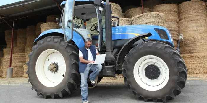 Ein Mann lehnt an einem blauen Traktor mit großen Reifen und schreibt etwas auf einem Laptop