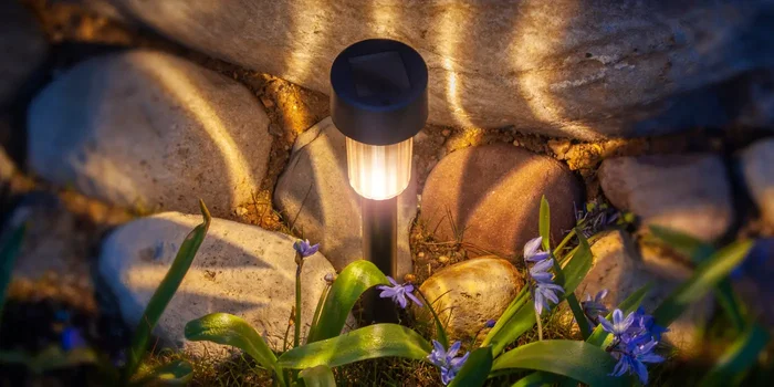 Eine Solarleuchte steht inmitten von Frühlingsblumen und wirft Lichtstreifen auf eine Steinformation